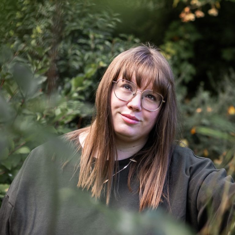 A young woman with long brown hair and glasses, standing among greenery.