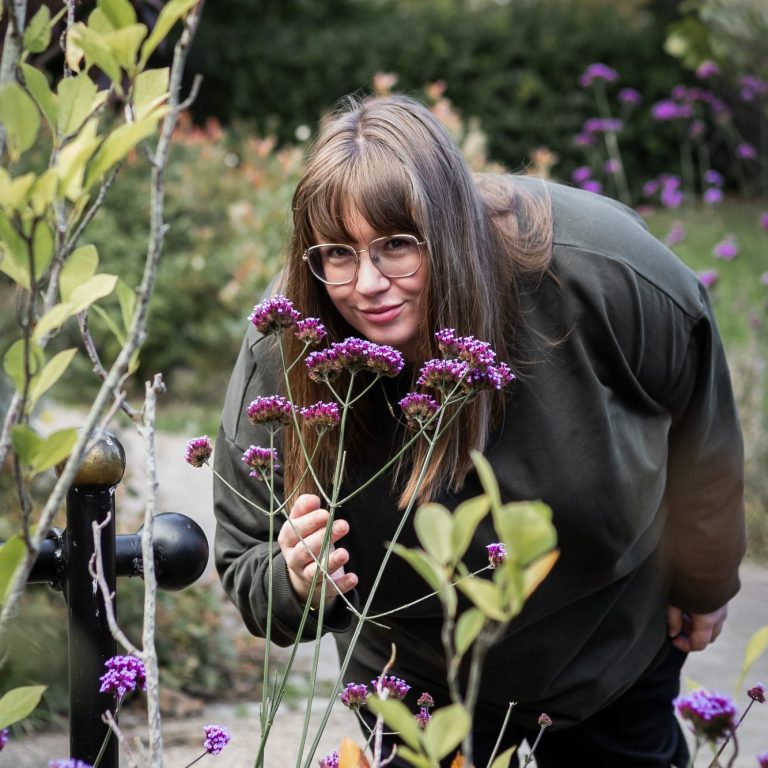 Rachel from Conductive Education with Rachel Short A person with glasses leans forward to smell flowers in a garden setting.